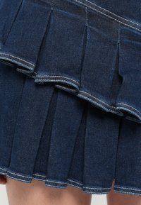 Tmavomodrá džínsová sukňa s vrstveným volánom, stredne dlhými plisovanými detailmi a kontrastným bielym prešívaním pozdĺž okrajov.