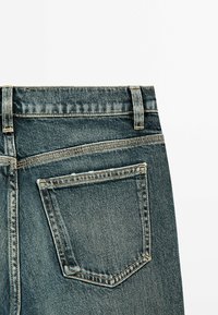 Poche arrière et ceinture d'un jean en denim bleu avec des coutures visibles et des passants pour ceinture sur fond blanc.