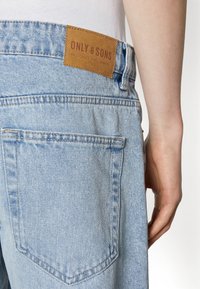 Light blue denim jeans with a leather patch labeled "ONLY & SONS" at the waist, featuring standard back pockets and a relaxed fit.