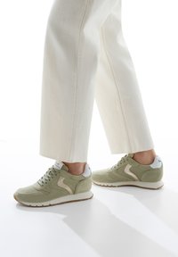 Grüne Wildledersneakers mit weißen Akzenten, ausgestattet mit einer strukturierten Oberfläche, gepolstertem Kragen und einer Sohle aus Naturkautschuk. Breite, gerade cremefarbene Hosen.