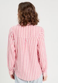 Chemise à manches longues avec des rayures verticales roses et blanches. Elle possède un col à rabat, un pli au dos et un ourlet arrondi. Texture de tissu douce.