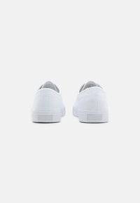 Zapatillas de lona blancas con un diseño de perfil bajo, parte superior texturizada y suelas de goma planas con un discreto detalle en el panel trasero.