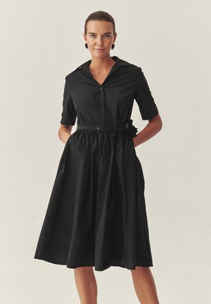 Zwarte shirtjurk met korte mouwen, knoopsluiting aan de voorkant, getailleerde taille en een bloemenaccent op de riem. Glad stof met een vloeiende rok.