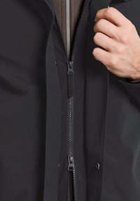 Svart ytterjacka med slät textur, med en central silverdragkedja och tryckknappar, delvis dragkedjad över ett brunt innerlager.