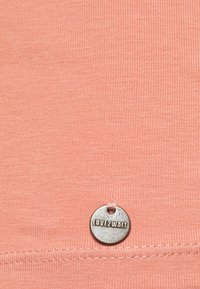 Tecido canelado de cor pêssego de uma camisa com uma etiqueta metálica com "LOVE2WAIT" em relevo, cosida na bainha. Textura suave.