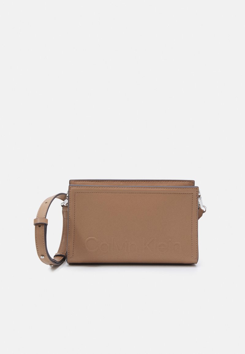 Tan krāsas ādas plecu soma ar iespiestu "Calvin Klein" logo, ar rāvējslēdzēja aizdari un regulējamu siksnu. Gluda tekstūra ar tīrām līnijām.