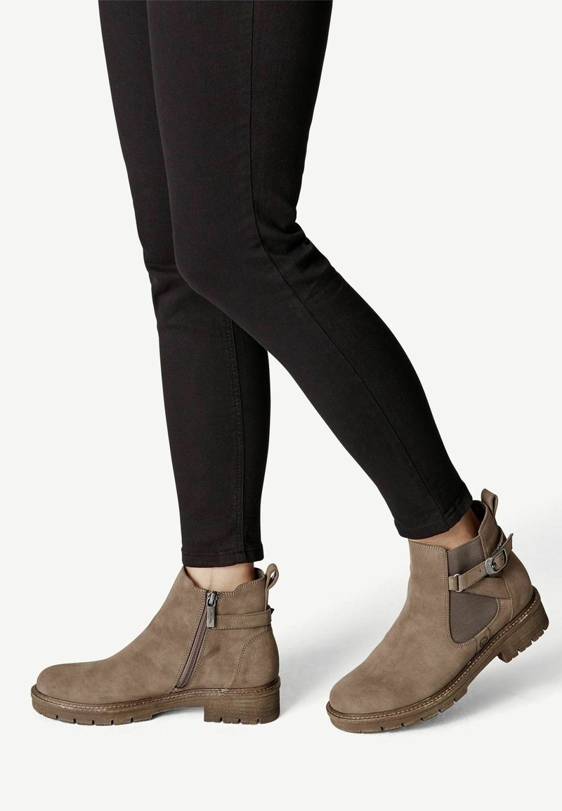 Jambes en pantalon noir portant des bottines en daim taupe avec fermeture éclair latérale, bride à boucle et semelle épaisse sur fond blanc.
