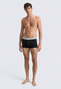 Fekete boxerrövidnadrágok fehér derékrésszel, amelyen a "TOM TAILOR" felirat olvasható, egy ing nélküli férfi modellön, szürke háttér előtt állva.