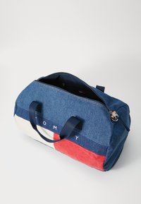 Modrá denimbová cestovná taška s červenými a krémovými akcentovými pruhmi, vybavená dvomi uchami a zipsovým uzáverom. Interiér je tmavý s detailnými vreckami.