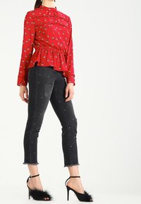 Röd blommig blus med peplummidja, långa ärmar och hög krage, som är kombinerad med svarta utsmyckade jeans och svarta klacksandaler.