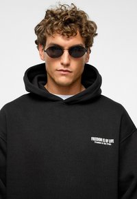 Zwarte hoodie met een kangoeroe zak, voorzien van witte tekst "VRIJHEID IS VAN LEVEN" op de linkerkant van de borst. Draagt een paar ovaalvormige zwarte zonnebril.