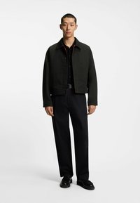 Musta lyhyt takki, jossa napitus, punaiset yksityiskohdat ja jämäkät hartiat, yhdistettynä mustiin housuihin ja mustiin kenkiin.