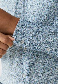 Ziedu apdruka uzkrekls ar zilu un baltu krāsu shēmu, pogu aprocēm un teksturētu audumu ar maziem, atkārtotiem modeļiem.