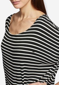 Camisa de manga larga de rayas blancas y negras con un corte holgado, cuello redondeado y pequeño logo cerca del dobladillo. Textura de tela suave.