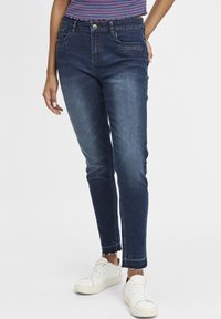 Jeans de denim azul oscuro con un ajuste slim, que cuentan con bolsillos frontales, un cierre de botones y detalles de dobladillo deshilachado. Llevados con zapatillas blancas.