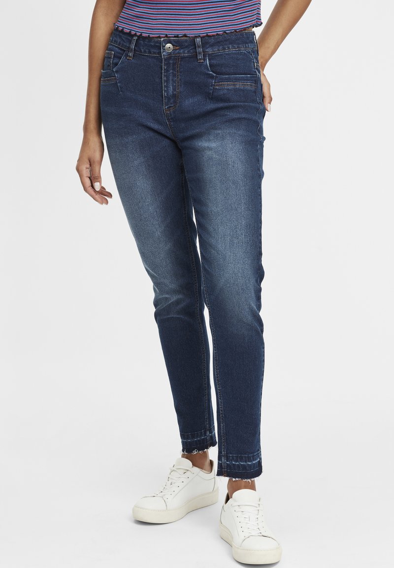 Jeans de denim azul oscuro con un ajuste slim, que cuentan con bolsillos frontales, un cierre de botones y detalles de dobladillo deshilachado. Llevados con zapatillas blancas.