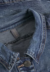 Jeansskjorta med klassisk krage, blå färg, synlig sömnad och en metallknapp med stjärndesign; etiketten står "CULTURE."