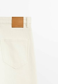 Jean en denim couleur crème avec une étiquette en cuir marron rectangulaire sur la taille, une poche arrière unique avec un détail cousu.