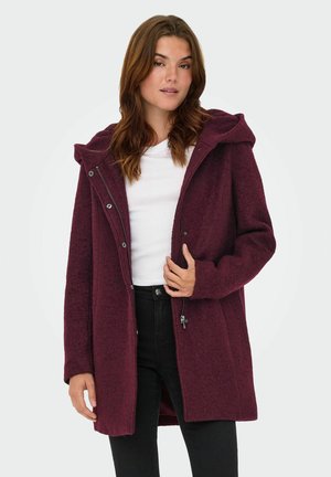 Manteau en mélange de laine bordeaux avec capuche, fermeture éclair à l'avant et boutons-pression. Présente un motif texturé et des poches latérales, porté sur une chemise blanche.