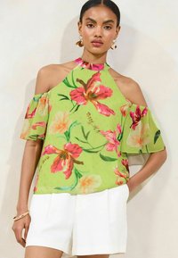 Blusa verde a fiori con spalle scoperte, decorata con fiori rosa e arancioni, un colletto arricciato e un tessuto leggero e testurizzato. Abbinata a pantaloni corti bianchi.