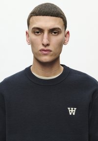 Marineblaues Sweatshirt mit Rundhalsausschnitt aus strukturiertem Stoff. Es hat ein kleines weißes Logo auf der linken Brust. Der Hintergrund ist hell und neutral.