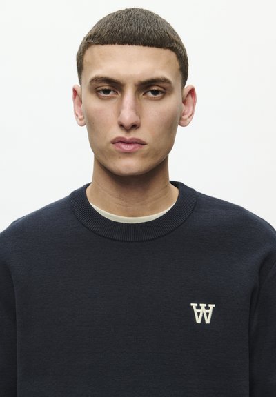 Sudadera de cuello redondo en color navy, hecha de un tejido texturizado. Presenta un pequeño logo blanco en el lado izquierdo del pecho. El fondo es claro y neutro.