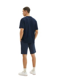 Marineblaues kurzärmliges T-Shirt in lockerer Passform, kombiniert mit marineblauen Shorts. Das Model trägt weiße Sneakers mit hellbeigen Socken. Das Design ist einfach gehalten.