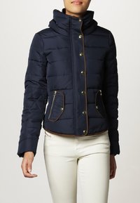 Marineblaue Daunenjacke mit hohem Kragen, goldenen Druckknöpfen, Reißverschlusstaschen und braunen Akzenten. Verfügt über ein gestepptes Design und Schulternähte.