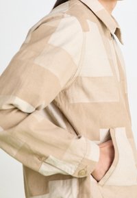 Beige vzorovaný kabát s límečkem, který má čtvercové nášivky v různých odstínech, boční kapsy a manžety na knoflíky.