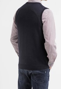 Marineblauer Strickpullover-Weste mit rundem Ausschnitt und geripptem Saum, getragen über einem gestreiften Hemd. Glatte Textur, figurbetontes Design.