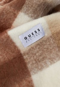 Brun og creme plaideret ulden tæppe med en struktureret overflade, der har et syet mærke med teksten "GUESS EST. 1981." Blødt og hyggeligt design.