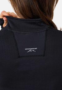 Musta urheiluyläosa, jossa on korkea kaulus, teksturoidusta kankaasta valmistettu, ja valkoinen logo vuorten kanssa selässä kaulan lähellä.