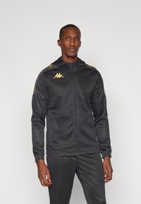 Black athletic jacket with hood, featuring gold logo and accents. Smooth fabric, front zipper, and side pockets. Designed for performance.