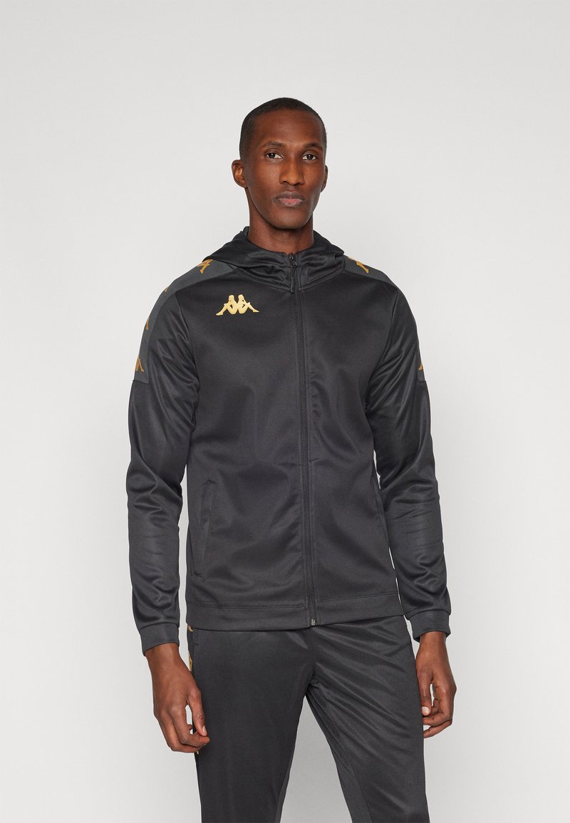 Zwarte sportjack met capuchon, voorzien van een gouden logo en accenten. Glad materiaal, voorrits en zijzakken. Ontworpen voor prestatie.