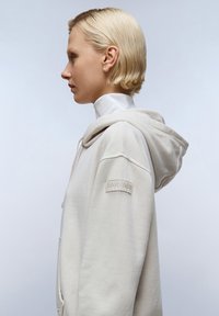 Beige hoodie met een trekkoord en een tekstuur logo patch op de linkermouw. Soepele stof en een relaxte pasvorm, met een zachte capuchon.