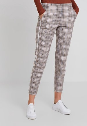 Personne portant un pantalon slim à carreaux avec des bandes blanches sur les côtés, mains dans les poches, associé à des baskets blanches et un haut à manches longues couleur rouille.
