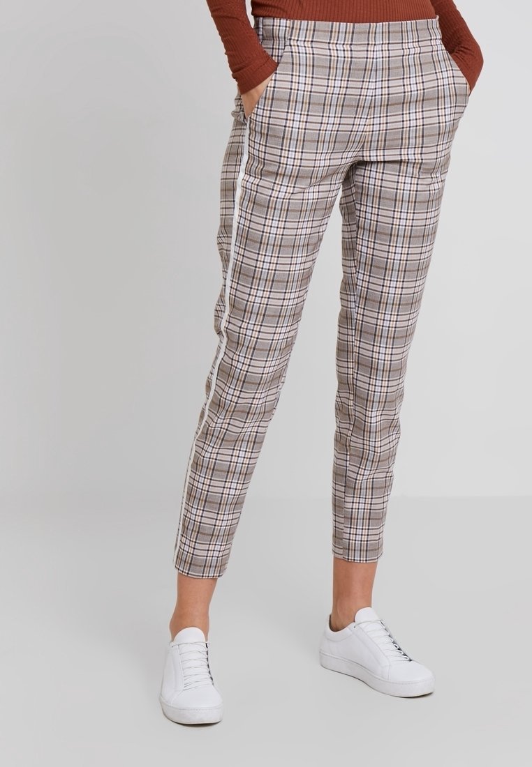 Personne portant un pantalon slim à carreaux avec des bandes blanches sur les côtés, mains dans les poches, associé à des baskets blanches et un haut à manches longues couleur rouille.