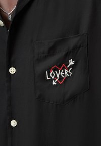 Czarna koszula z krótkim rękawem z kieszonką na piersi oraz haftowanym wzorem z czerwonym sercem, białym napisem "LOVERS" i elementami strzały.