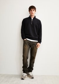 Schwarzer Zip-Hoodie mit hohem Kragen, kombiniert mit ausgewaschenen, dunklen Jeans und grauen Sneakers. Glatter Stoff, lockere Passform, standardmäßige Nähdetails.