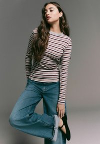 Randig långärmad topp i grått, rosa och beige; högmidjade, vida blå jeans; svarta skor; modellen poserar med ett ben böjt.