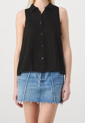 Femme portant une chemise noire sans manches boutonnée et une jupe en jean bleu avec des coutures verticales et un ourlet effiloché, debout devant un fond uni.