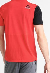 Camiseta atlética de manga corta en rojo y negro con una ligera textura, que presenta un logo de Reebok en la parte superior de la espalda. Combinada con pantalones cortos grises.