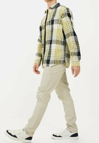 Camicia a quadri gialla e nera con due tasche sul petto, abbinata a pantaloni beige e sneaker bianche con dettagli neri. Modello in posa mentre cammina.
