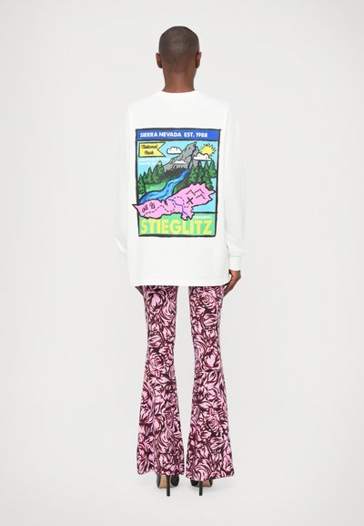 Cămașă albă cu mânecă lungă, cu un grafic colorat de peisaje și text pe spate, asociată cu pantaloni evazați cu model floral roz.
