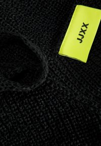 Tela negra texturizada con un patrón tejido, que presenta una etiqueta amarilla brillante marcada con "JJXX". La tela tiene una superficie táctil y elevada.