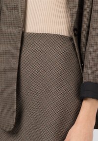 Bruine geruite blazer met een subtiele textuur, gecombineerd met een geribde lichtbeige top. Manchet van de mouw heeft een zwarte randdetail.