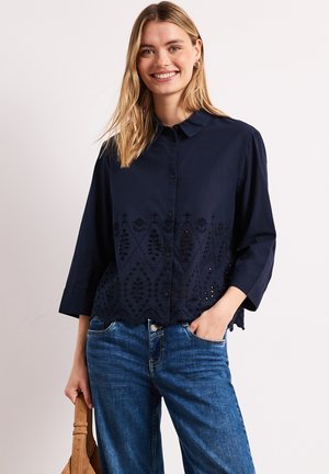 Donna sorridente con capelli biondi che indossa una camicetta blu navy con bottoni e motivo traforato e jeans blu, tiene una borsa beige in una mano.