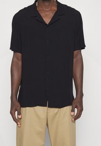 Camicia nera a maniche corte con bottoni, colletto con scollo a lancia, tessuto liscio e vestibilità comoda, indossata sopra pantaloni beige.