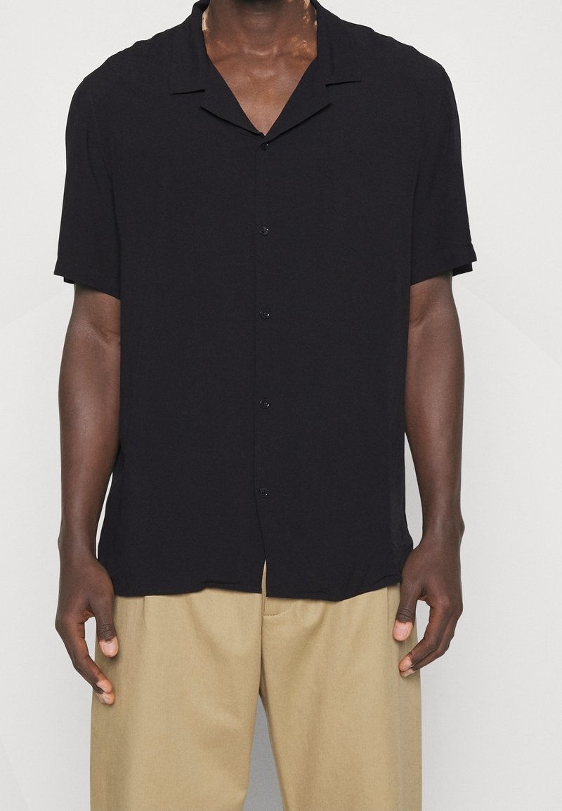 Camicia nera a maniche corte con bottoni, colletto con scollo a lancia, tessuto liscio e vestibilità comoda, indossata sopra pantaloni beige.