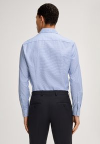 Camicia azzurra a fantasia con colletto stretto e maniche lunghe, abbinata a pantaloni scuri con due tasche posteriori.